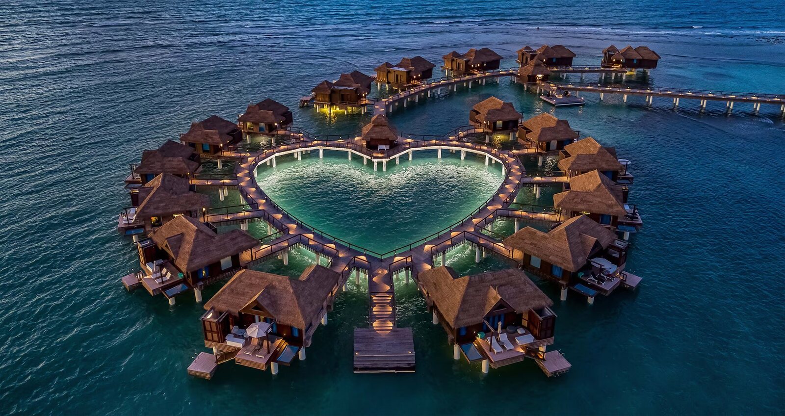 Heart-shaped ring of overwater villas lit at dusk at Sandals Caribbean Cay, Jamaica, booked by a Sandals travel agent